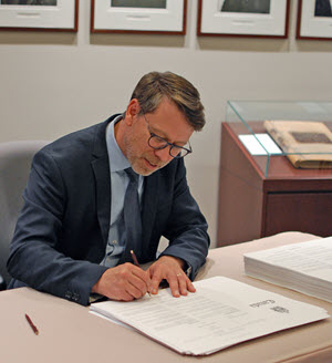 Un homme d'âge moyen portant des lunettes assis derrière un bureau signe une pile de documents d'allure officielle.