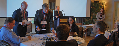 Six personnes en tenue de ville examinent des documents tout en discutant, rassemblés autour d'une table ronde dans une salle de réunion. Une présentation est affichée sur deux grands écrans dans le fond de la salle