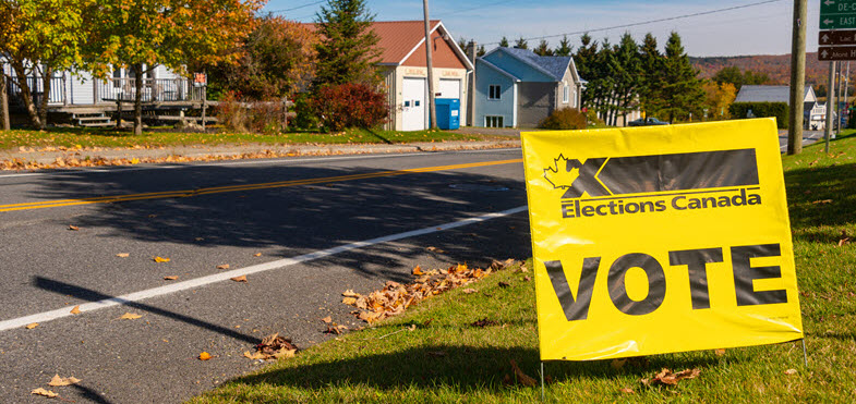 Un panneau jaune portant le mot 'vote' et le logo d'Élections Canada est placé sur un bout de pelouse devant une rue vide d'un quartier rural