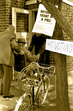 Black-and-white photo of a man in a wheelchair being assisted into a polling station during the 1963 General Election. The photo includes the posted Notice of Grant of a Poll on a tree outside the polling station.