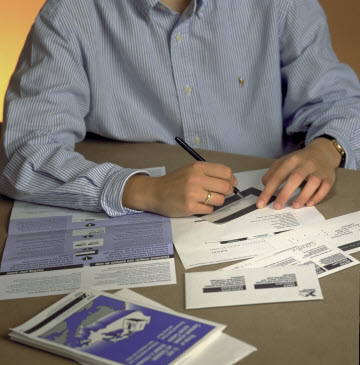 Photo of a person's hands using a pen to fill out a special ballot. Papers with instructions and a pair of envelopes sit on the table near the ballot. 