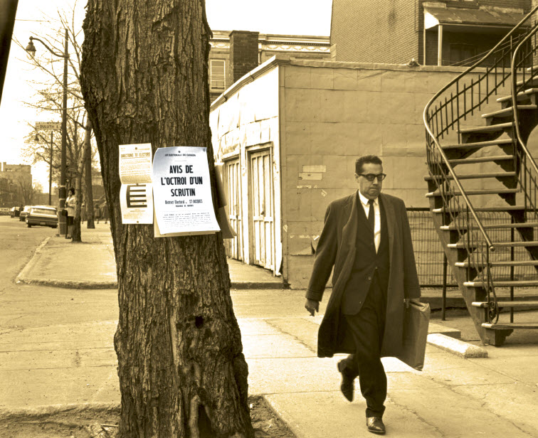 Photo en noir et blanc d'un homme à pied, en complet et manteau long, qui passe près d'un arbre sur lequel sont fixés des documents, dont un où on lit « Notice of Election » [avis d'élection].