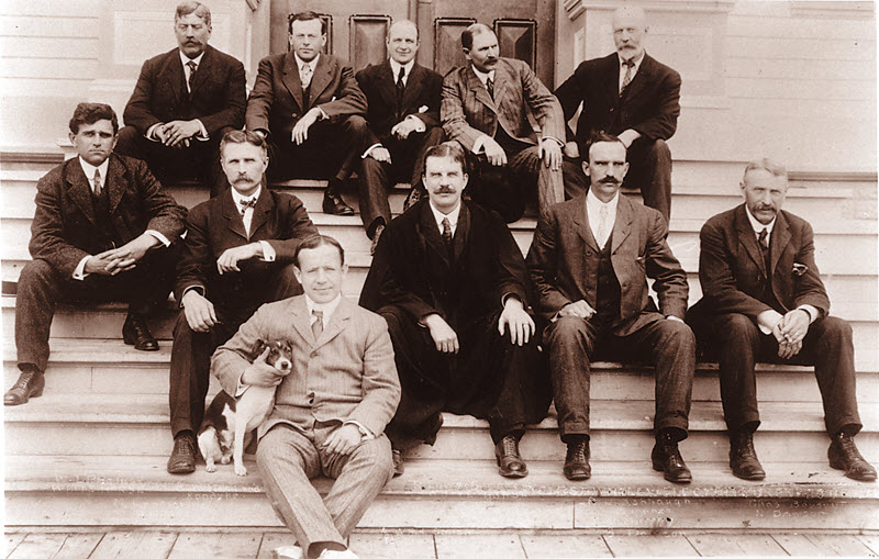Black and white photo of a group of men seated on the steps of a building.