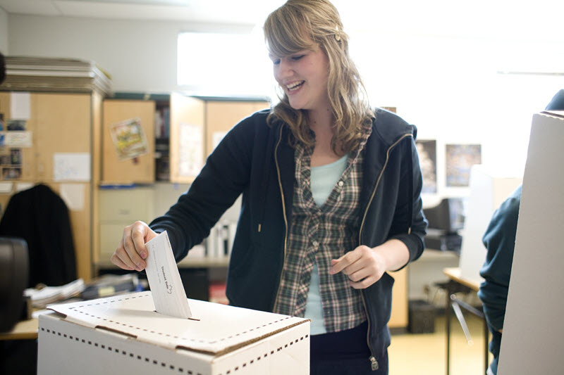 Photo showing a teenage girl in a classroom putting a ballot into a ballot box.