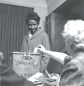 Black-and-white photo showing a Black woman watching the deputy returning officer placing her ballot in the ballot box on voting day in the 1963 federal election.