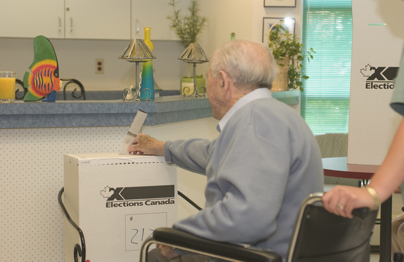 Photo d'un homme âgé en fauteuil roulant déposant un bulletin de vote dans une urne en carton d'Élections Canada installée dans une cuisine.