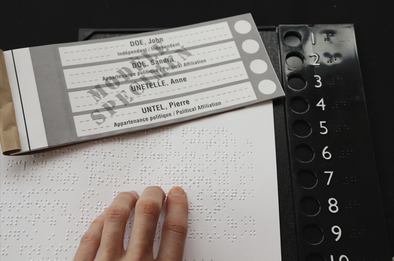 Photo d'un bulletin de vote, d'une liste des candidats en braille et d'un gabarit de vote permettant de maintenir le bulletin en place. Le gabarit comporte des chiffres en relief et en braille.