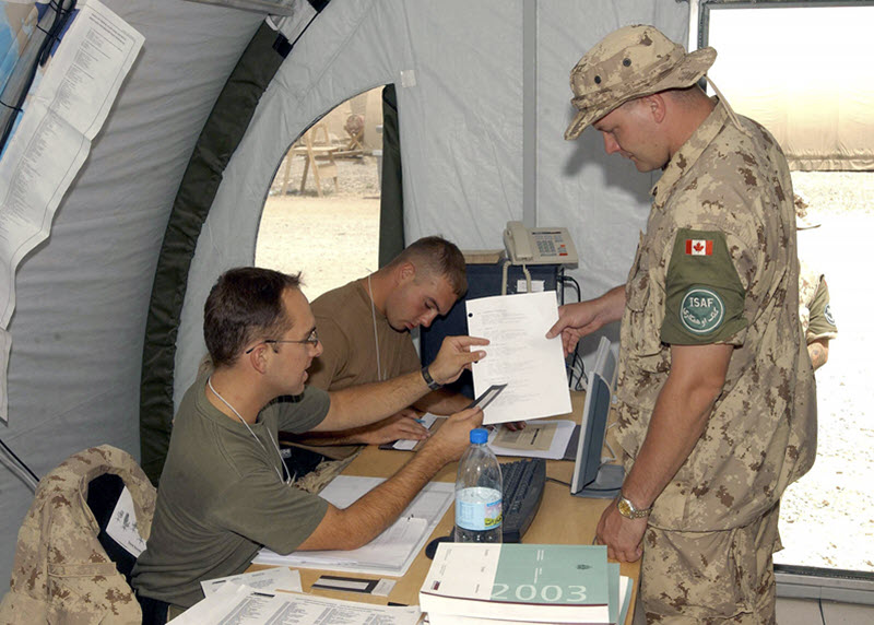 Photo of a soldier dressed in a camouflage uniform standing at a polling station and looking at a document that an election worker seated behind the table holds up and points to.