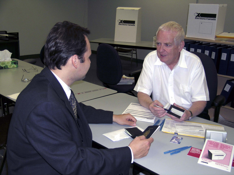 Photo of a seated man holding a piece of identification and speaking to an election worker who is holding an election ballot.