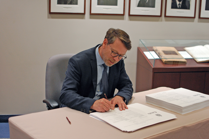Photo of the Chief Electoral Officer, Stéphane Perrault, seated at a desk signing a large document. Next to him is a stack of similar documents.