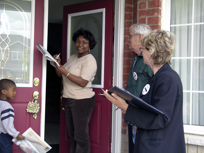 A woman stands in the open doorway of a home as she signs a document. Two Elections Canada workers holding clipboards look on as they stand across from her.