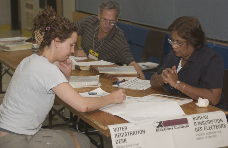 Photo of a woman sitting at a table marked 'Voter Registration Desk' and signing a piece of paper in front of two election workers who are seated across from her.