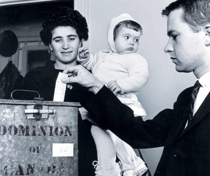 Photo of a woman holding a baby as she watches a man puts a paper ballot into a metal ballot box.