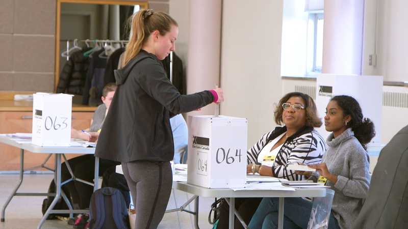 Photo de deux agents électoraux assis à une table, regardant une jeune femme déposer son bulletin de vote dans une urne.